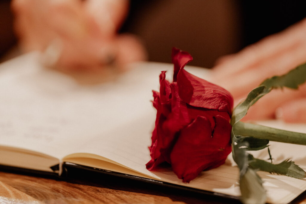 Eine rote Rose liegt auf einem aufgeschlagenen Notizbuch, in das gerade geschrieben wird